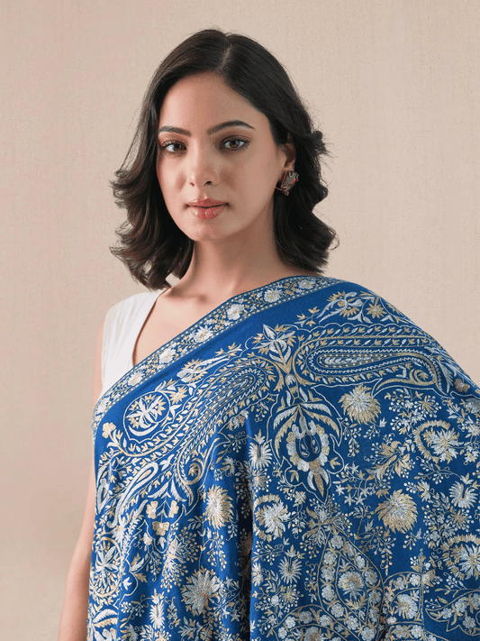 A woman wearing a royal blue pashmina shawl with intricate gold and silver zari embroidery in floral and paisley patterns.