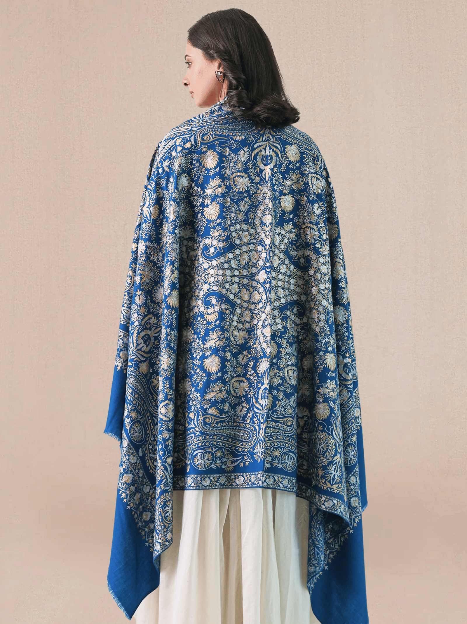 A woman wearing a royal blue pashmina shawl with intricate gold and silver zari embroidery in floral and paisley motifs, and a floral border.