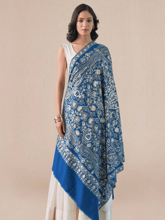 A woman wearing a royal blue pashmina shawl with intricate gold and silver zari embroidery in floral and paisley motifs, and a floral border.